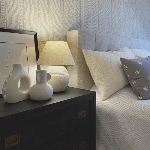 ANNA’S DREAM glowne w stylu justyna sonta sypialnia Wnętrze sypialni w przytulnym stylu. Widok na ścianę z tapetą w delikatny wzór, szafkę nocna w kolorze szaro-czarnego drewna. Na stoliku stoi ceramiczna lampa z lnianym kloszem i grafiką z obrazkiem kobiety - Warszawa W stylu Justyny