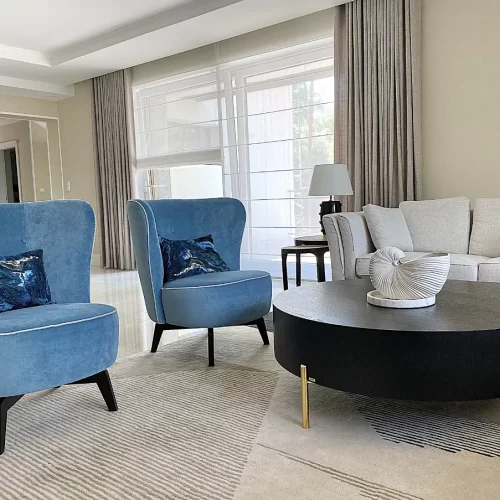 CONTEMPORARY ELEGANCE glowne w stylu justyna sonta salon.2 Widok na salon w stylu Contemporary Elegance z dwoma fotelami w kolorze błękitnym, czarny stolik kawowy na złotych metalowych nóżkach, beżowe ściany i fragment sofy w szaro-beżowej tapicerce - Warszawa W Stylu Justyny